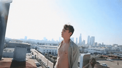 A Shirtless Man Is Standing On A Rooftop Overlooking A City Skyline . GIF