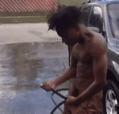 A Shirtless Man Is Washing A Car With A Hose In A Driveway . GIF
