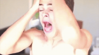 A Shirtless Man Is Yawning With His Mouth Open While Holding His Head In Front Of A Hair Dryer . GIF