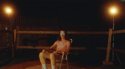 A Shirtless Man Sits In A Chair In Front Of A Wooden Fence At Night GIF