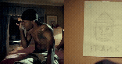 A Shirtless Man Sits On A Bed Next To A Framed Drawing Of Him GIF