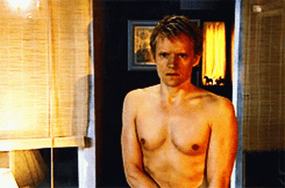 A Shirtless Man Stands In A Bathroom With A Picture On The Wall Behind Him GIF
