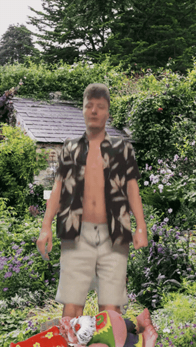 A Shirtless Man Stands In A Garden With Flowers GIF
