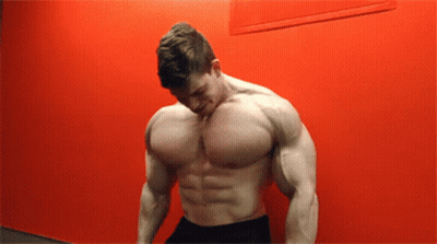 A Shirtless Man Stands In Front Of A Red Wall With His Head Down GIF