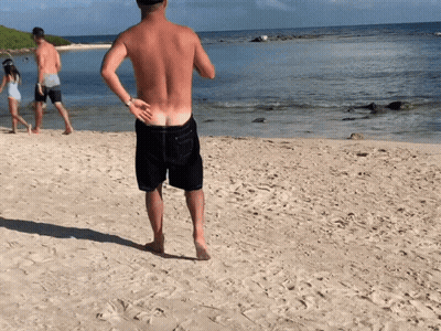 A Shirtless Man Stands On A Sandy Beach Looking At The Ocean GIF