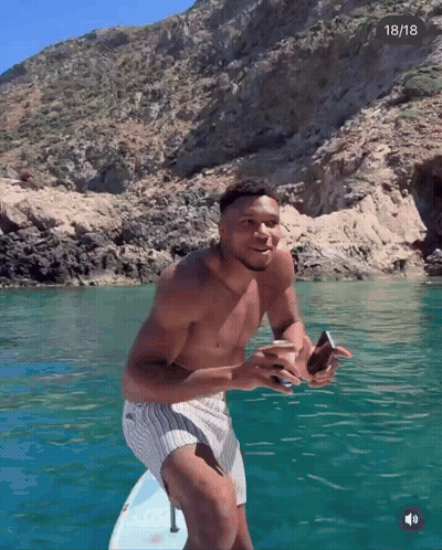 A Shirtless Man Stands On A Surfboard In The Water With The Date 18/18 In The Corner GIF