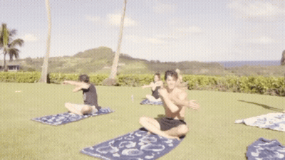 A Shirtless Man Stretches His Arms While A Woman Sits On The Grass GIF
