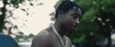 A Shirtless Man Wearing A Chain Around His Neck Is Standing In Front Of Trees . GIF