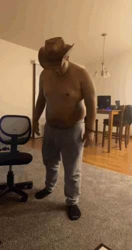 A Shirtless Man Wearing A Cowboy Hat And Sweatpants Stands In A Living Room GIF