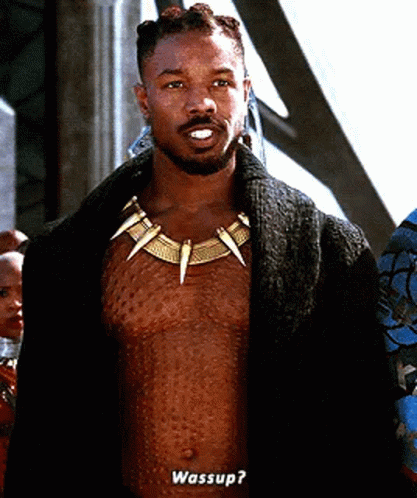 A Shirtless Man Wearing A Necklace With Sharp Teeth Is Standing In Front Of A Group Of People And Talking . GIF