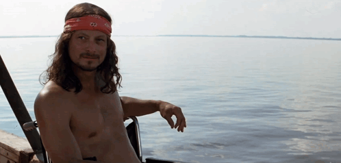 A Shirtless Man Wearing A Red Bandana With The Letters Co On It Sits In Front Of A Body Of Water GIF