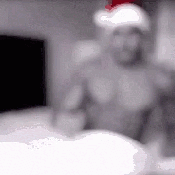 A Shirtless Man Wearing A Santa Hat Is Standing In A Dark Room GIF