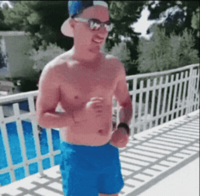 A Shirtless Man Wearing Blue Shorts And A Hat Is Standing On A Balcony . GIF