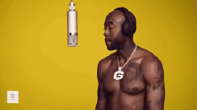 A Shirtless Man Wearing Headphones And A Necklace With A G On It Is Standing In Front Of A Microphone . GIF