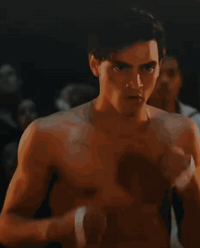 A Shirtless Man With A Bandage On His Wrist Is Standing In A Dark Room With Other Men . GIF