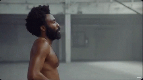 A Shirtless Man With A Beard And Afro Is Standing In A Warehouse . GIF