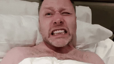 A Shirtless Man With A Beard Is Making A Funny Face While Laying In Bed GIF