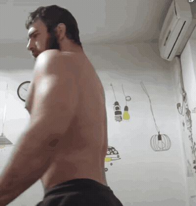 a shirtless man with a beard is standing in a room with a clock on the wall .