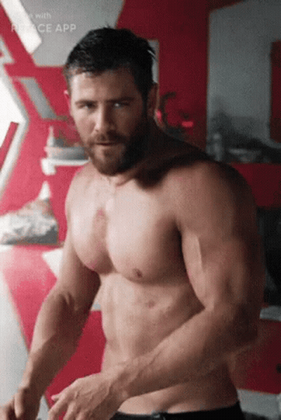 A Shirtless Man With A Beard Is Standing In Front Of A Red Wall In A Room . GIF
