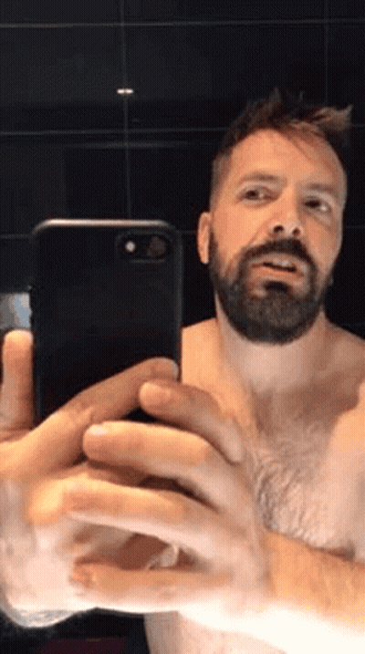 A Shirtless Man With A Beard Is Taking A Selfie With His Cell Phone In A Bathroom . GIF