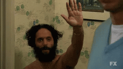A Shirtless Man With A Beard Is Waving His Hand In Front Of A Wall . GIF