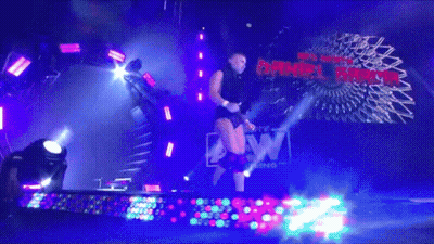 A Shirtless Man With A Black Scarf Around His Neck Stands In A Dark Room GIF
