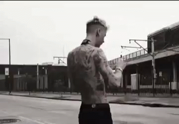 A Shirtless Man With A Lot Of Tattoos Is Standing On A Street In Front Of A Building . GIF