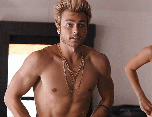 A Shirtless Man With A Necklace Around His Neck Stands In Front Of A Mirror GIF