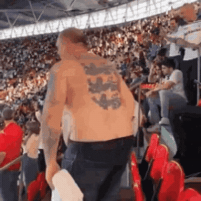 A Shirtless Man With A Tattoo On His Back Is Standing In Front Of A Crowd Of People . GIF