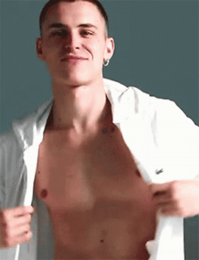 A Shirtless Man With A Tattoo On His Chest Is Taking Off His Shirt And Smiling . GIF