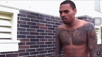 A Shirtless Man With A Tattoo On His Chest Stands In Front Of A Brick Wall GIF