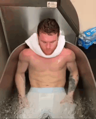 A Shirtless Man With A Towel Around His Neck Is Sitting In A Tub Of Water . GIF