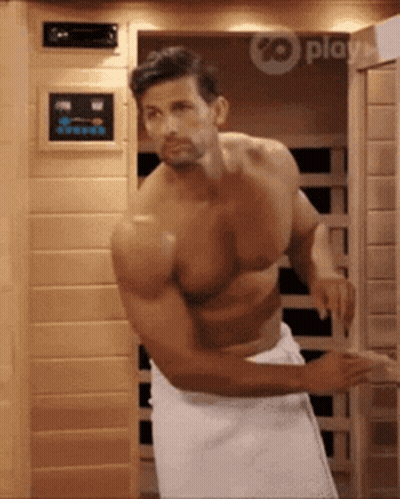 A Shirtless Man With A Towel Around His Waist Is Standing In Front Of An Infrared Sauna . GIF