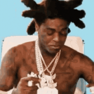 A Shirtless Man With Dreadlocks And A Necklace Is Sitting In A Chair Eating A Piece Of Food . GIF
