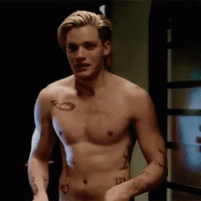 A Shirtless Man With Tattoos On His Arm And Chest Is Standing In Front Of A Door . GIF