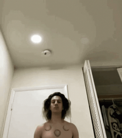 A Shirtless Man With Tattoos On His Chest Is Standing In Front Of A Door In A Bathroom . GIF