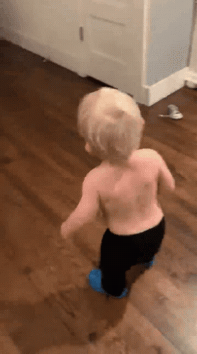 A Shirtless Toddler Is Walking On A Wooden Floor . GIF