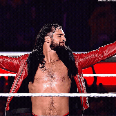 A Shirtless Wrestler In A Red Jacket Stands In A Wrestling Ring With His Arms Outstretched GIF