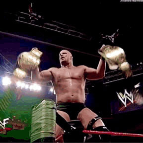 A Shirtless Wrestler Is Holding Up His Championship Belts In A Wrestling Ring GIF