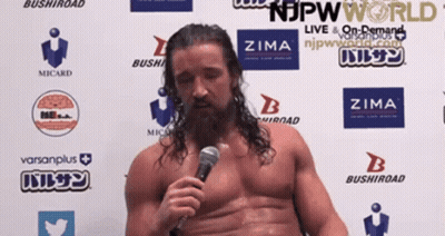 A Shirtless Wrestler Is Speaking Into A Microphone In Front Of A Wall With Logos For Njpw World And Zima GIF