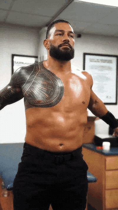 A Shirtless Wrestler With A Beard And Tattoos On His Chest Is Standing In A Room . GIF