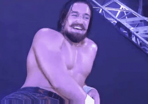 A Shirtless Wrestler With A Beard Is Smiling While Standing In Front Of A Purple Wall . GIF