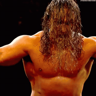 A Shirtless Wrestler With Long Hair Is Standing In The Dark . GIF