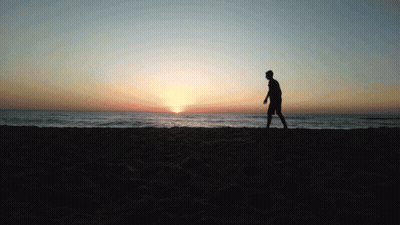 A Silhouette Of A Man Walking On A Beach At Sunset GIF