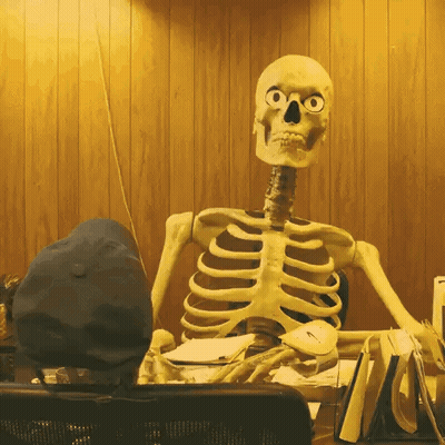 A Skeleton Is Sitting At A Desk With A Hat On GIF