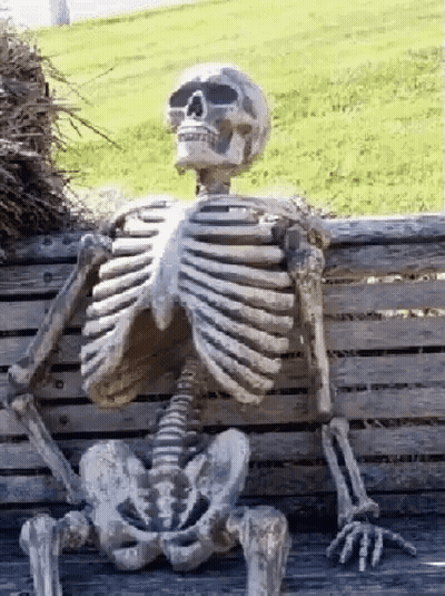 A Skeleton Is Sitting On A Wooden Bench In The Grass . GIF