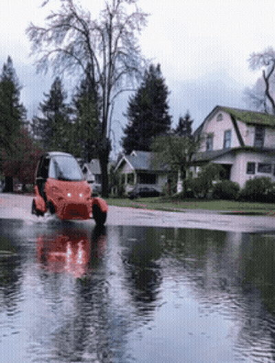 A Small Orange Vehicle Is Driving Through A Flooded Street GIF