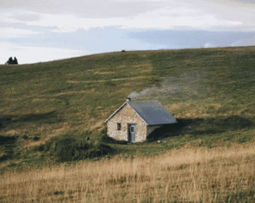 A Small Stone House Sits On Top Of A Grassy Hillside GIF