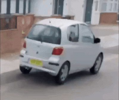 A Small White Car Is Driving Down A Street Next To A House . GIF