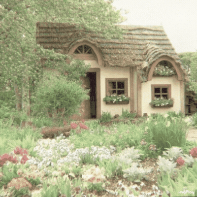 A Small White House With A Thatched Roof Is Surrounded By Flowers And Trees . GIF